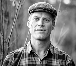 black and white portrait of Basil Camu in a plaid shirt and tweed cap, surrounded by tree branches