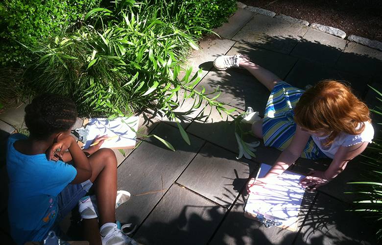 two children sitting on a stone path trace shadows from plants onto paper