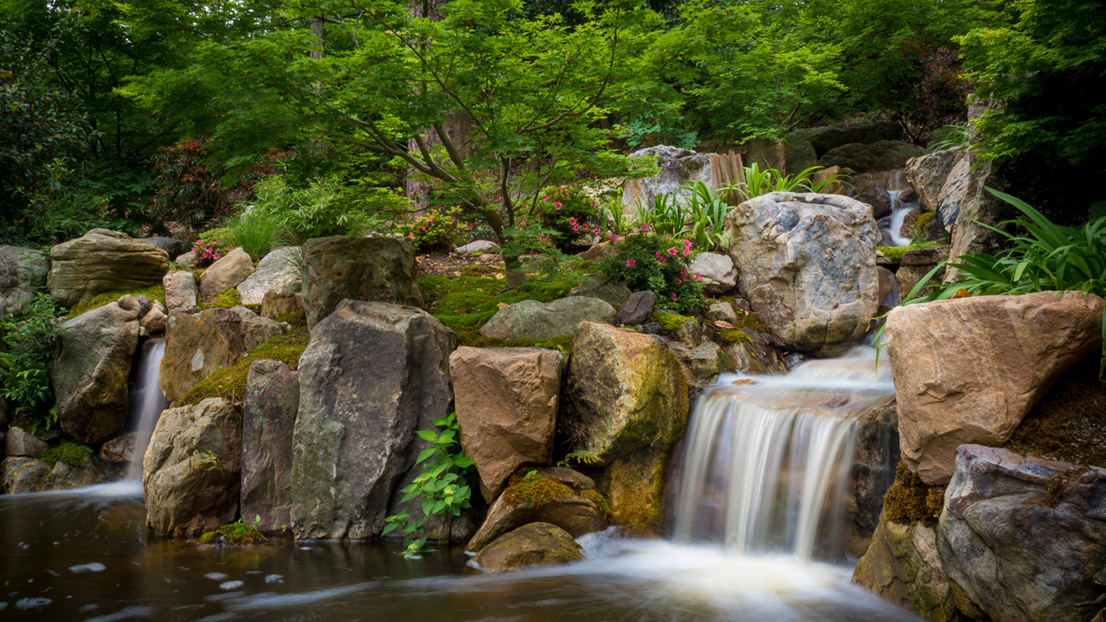 A small waterfall flows over a group of large boulders placed artistically in a garden space with a small Japanese maple tree and petite pink flowers above the boulders