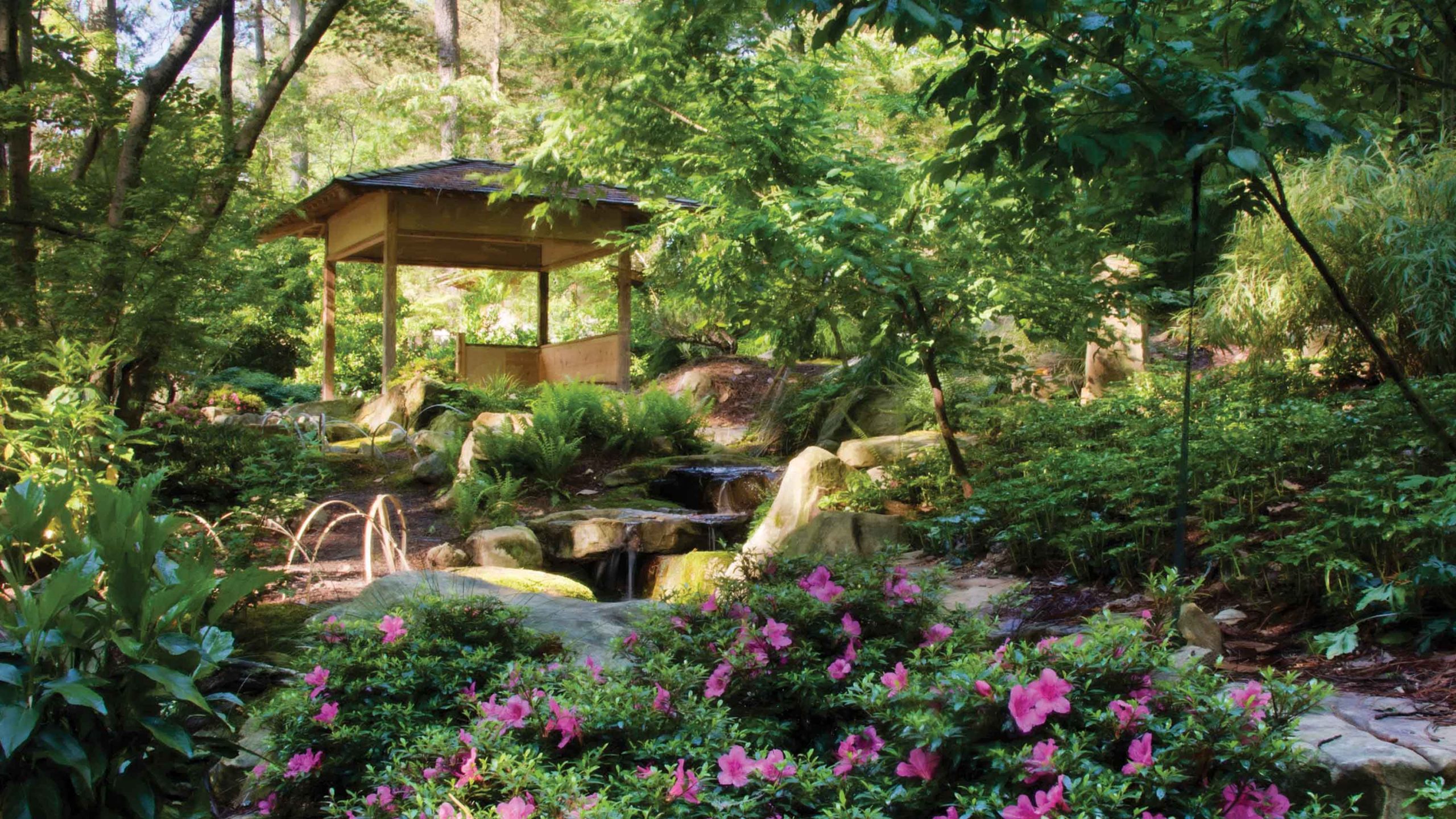 Wooden gazebo on a hill with green trees and shrubs, boulders and pink flowers in the foreground.