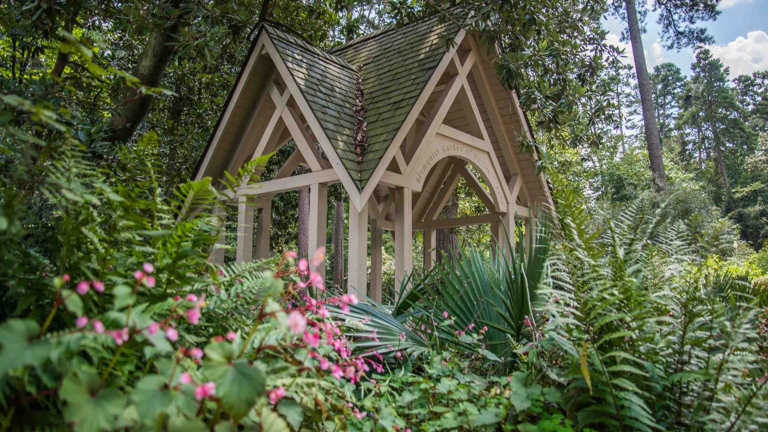 A wooden garden gatehouse with pink flowering shrubs around it.