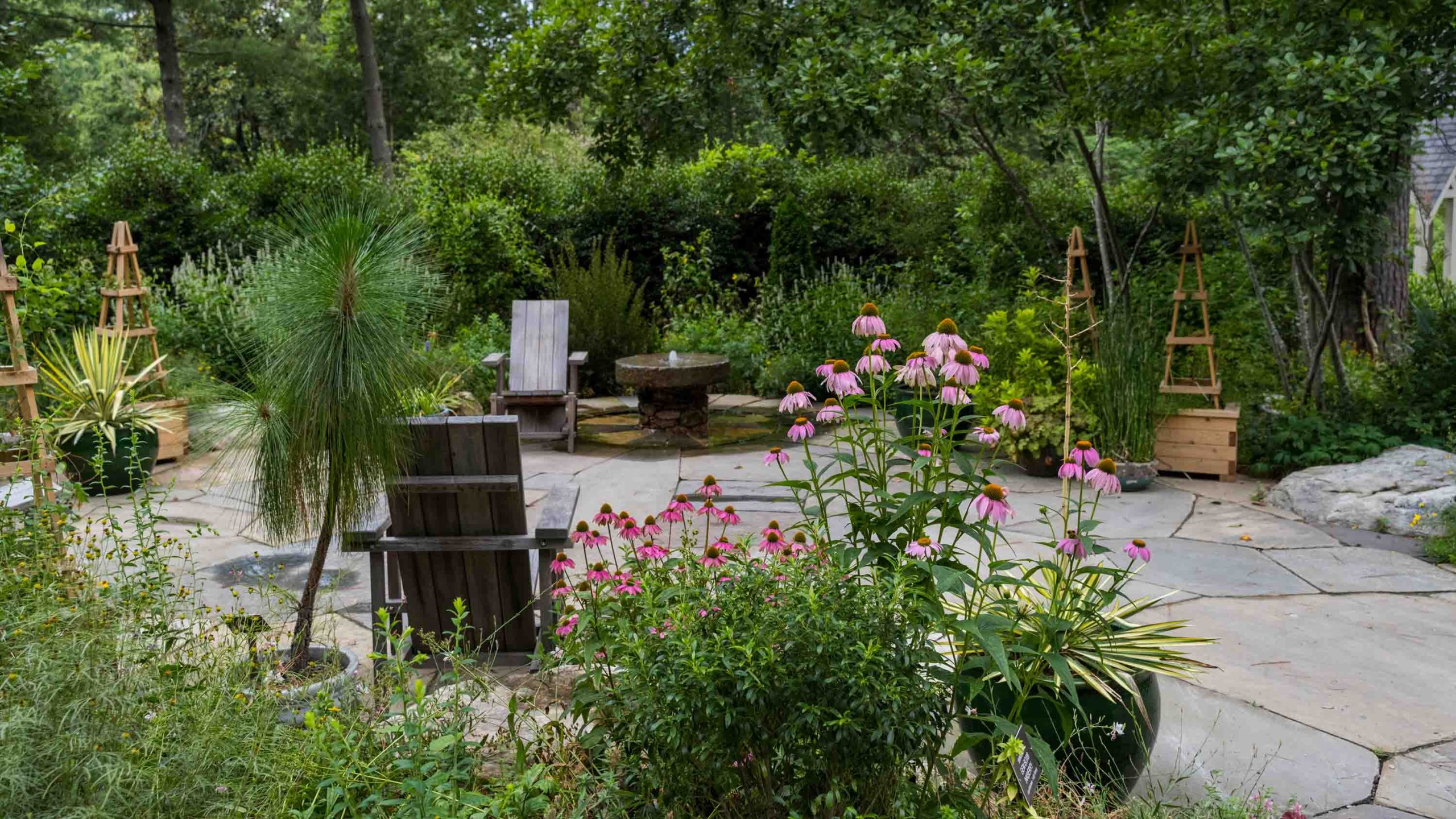 A stone garden patio with two Adirondack chairs, a wooden trellis with climbing clematis flowers and a small stone garden fountain in the distance.