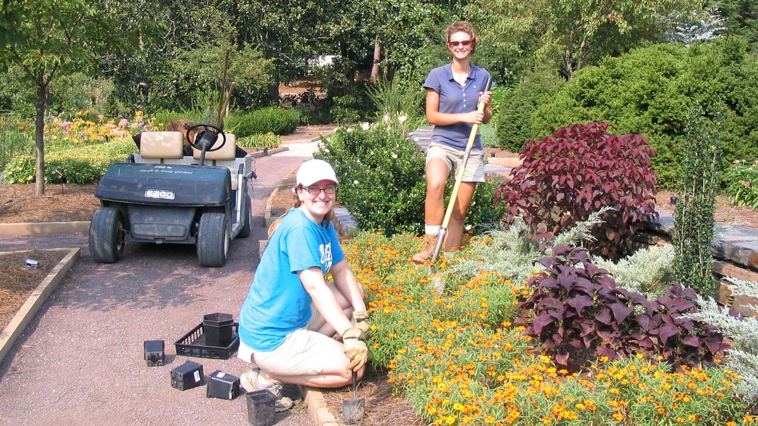 Two people in a colorful garden setting, surrounded by orange flowers, one kneeling next to empty plant pots, one standing holding a shovel. Behind them is a golf cart on a gravel path.