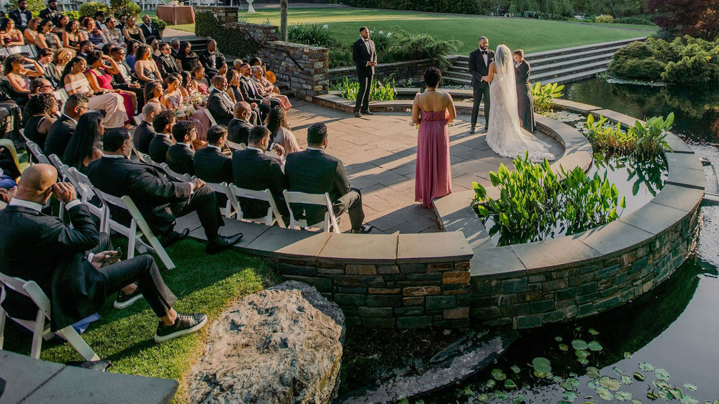 A large crowd of people in outdoor amphitheater facing a wedding ceremony at the edge of a pond, with trees and an arbor in the distance.