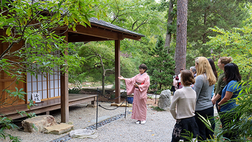 A woman in a kimono points out features of the Japanese teahouse to a group of bystanders