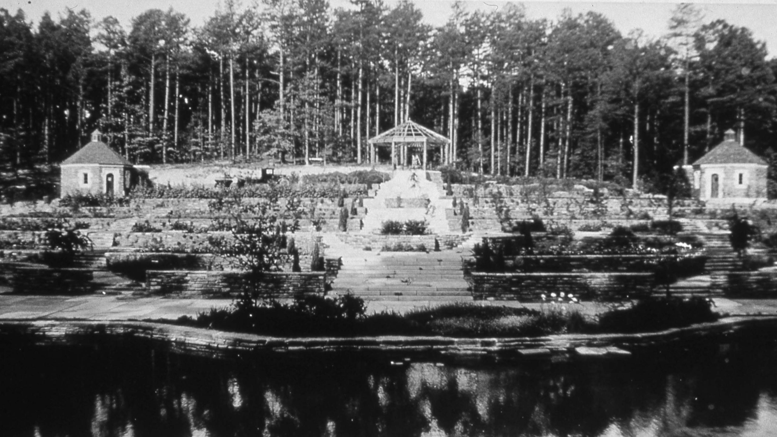 a black and white archival photo of a garden with a metal pergola structure at top and reflective pond at bottom.