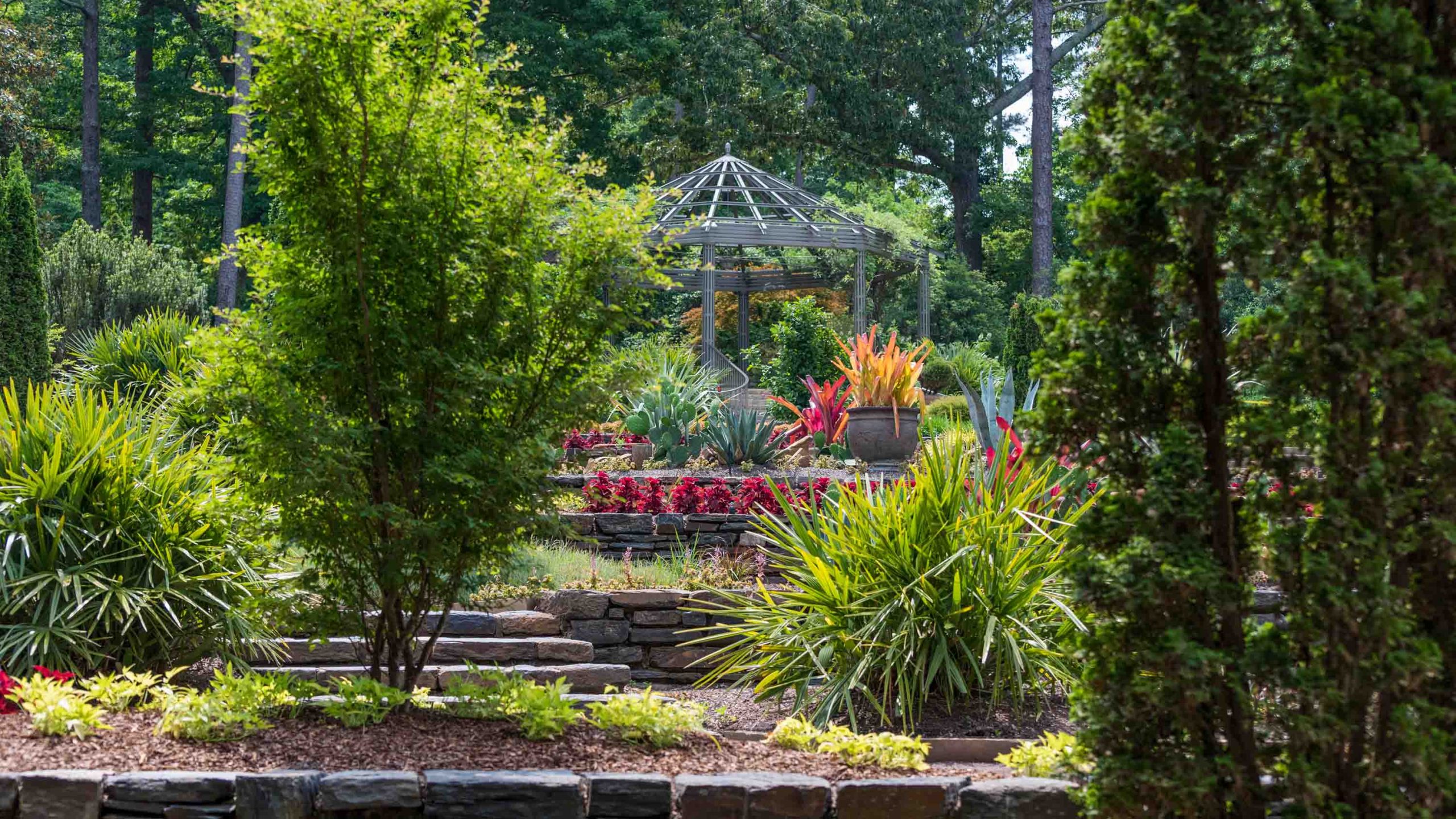 A metal pergola surrounded by brightly colored potted plants and trees.